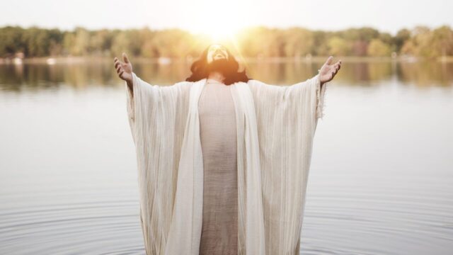 A vertical shot of the Jesus Christ standing in the water with his hands and head towards the sky