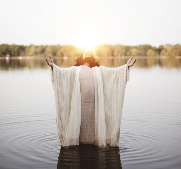 A vertical shot of the Jesus Christ standing in the water with his hands and head towards the sky