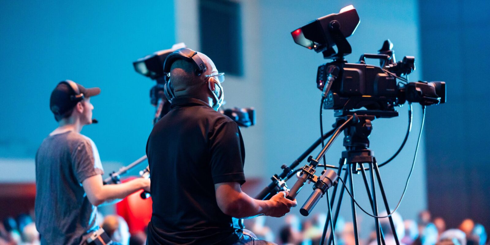 Two cameramen operate professional video cameras at an indoor event with a lively audience.